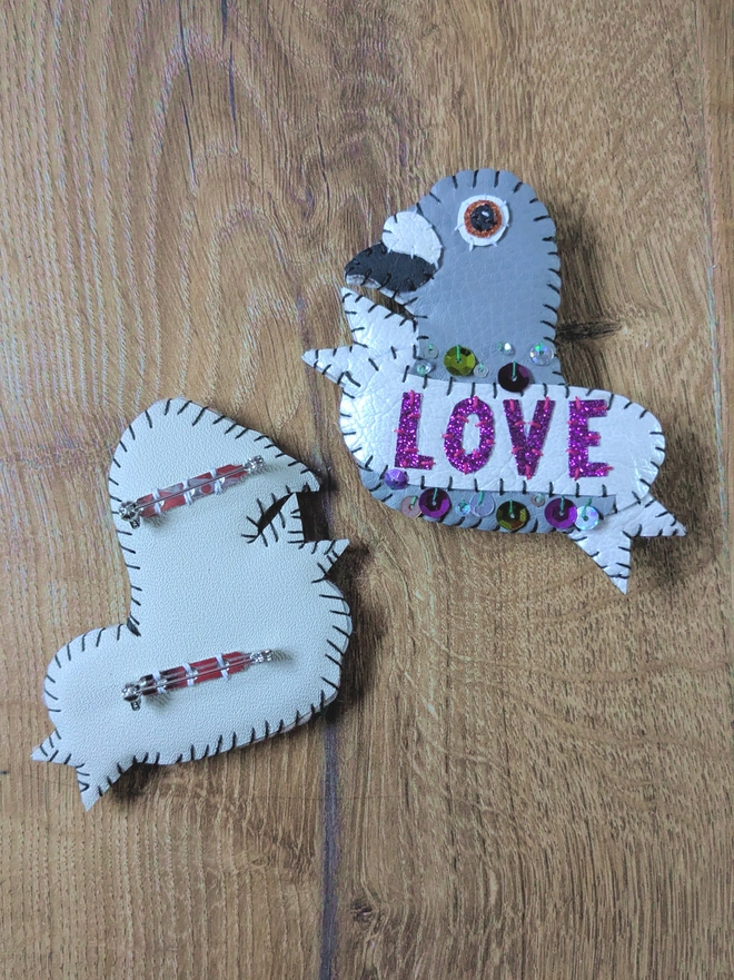 Front and back view of a hand-stitched white and grey faux leather pigeon brooch, personalised with purple glitter ‘LOVE’ lettering and colourful sequins, with two secure brooch fastenings on the reverse.
