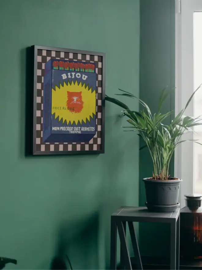 An illustrated vintage-style matchbox labeled “BIJOU.” The box is dark blue with a jagged yellow burst shape in the center, featuring a bright red cat’s face. Text on the burst reads “PRICE Re. 0.06.” Along the bottom, white lettering says “MON PRECIEUX CHAT ALLUMETTES TRIOMPHE.” The top of the matchbox shows red-tipped matches with green stems. The background is a checkered pattern in black and white.