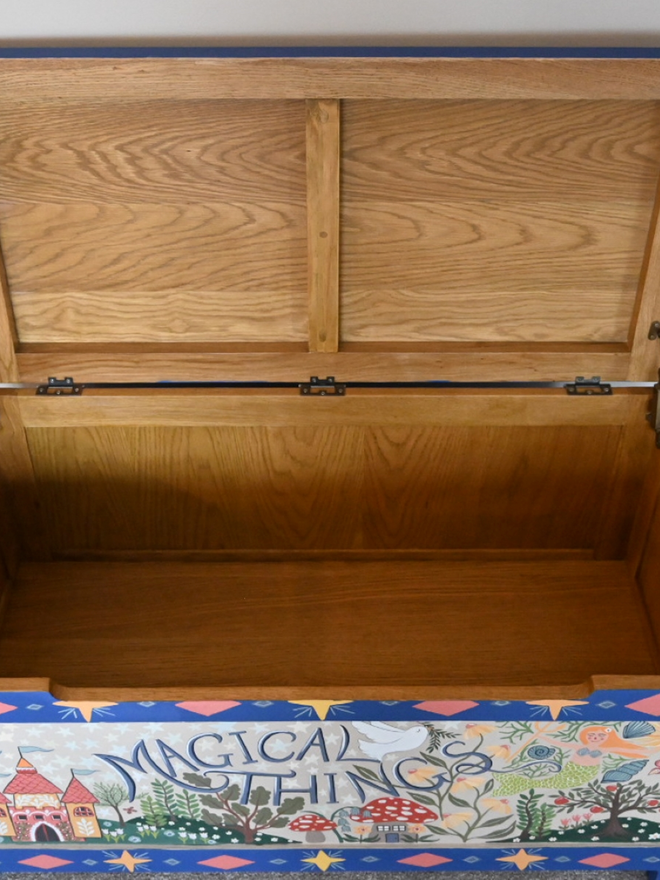 A straight-on view into an open wooden chest. The interior is a light, natural wood color with a smooth finish. The exterior of the chest is painted a vibrant blue and features the phrase "Magical Things" in a playful font, centered below the lid. Beneath the phrase is a colorful scene with whimsical illustrations, including a pink castle, trees, mushrooms, and other fairytale-inspired elements. The inside of the open lid is the same natural wood as the chest's interior. The chest is open and empty, suggesting readiness for storage or play.