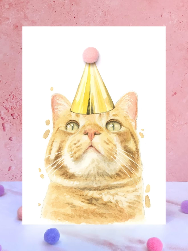 A greeting card featuring a hand painted design of a ginger tabby cat, stood upright on a marble surface surrounded by pompoms. 