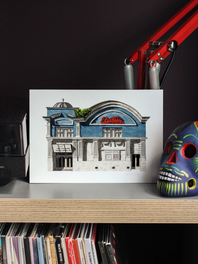 An architectural illustration of the Electric Cinema on a print, displayed on a shelf above vinyl records alongside a red desk lamp and a colorful decorative skull.