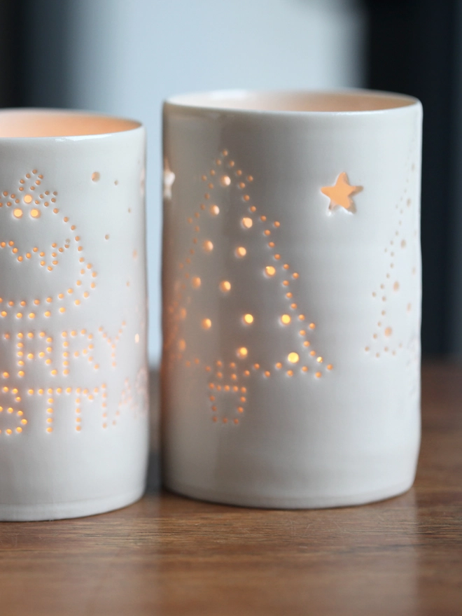 A porcelain tealight holder, lit from inside glows on a tabletop. The tealight holder is pierced with tiny holes that allow the light to come through, in the shape of Christmas trees and stars