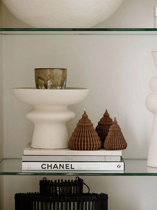 Brown Origami Christmas Tree Warm Candles (Set Of 2). Three brown fluted cone tree candles in graduating sizes, arranged on a stack of books beside a cream pedestal bowl and ceramic cup. The small sculptural candles add earthy texture and festive charm to a styled shelf.