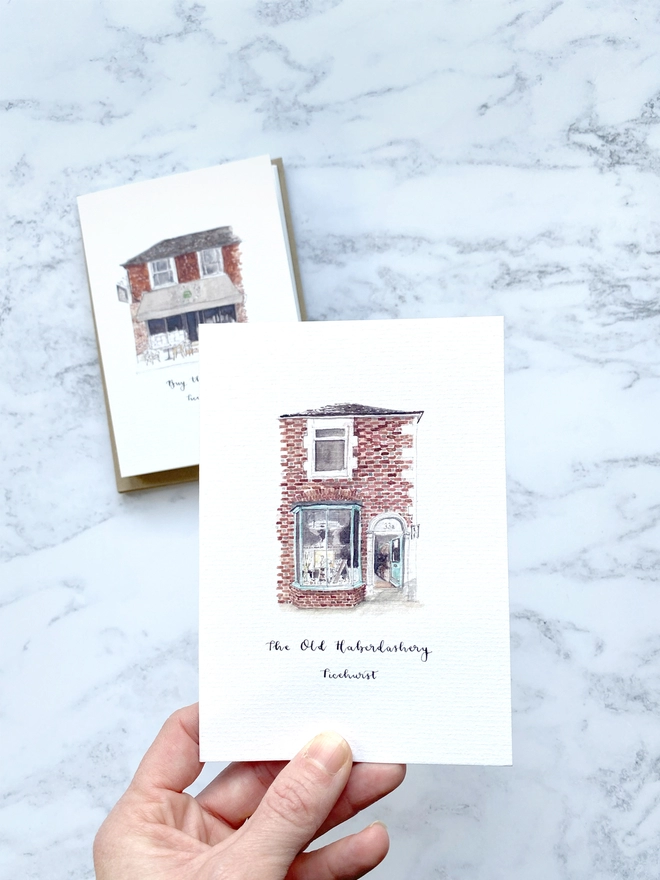 Greetings card showing watercolour painting of The Old Haberdashery, haberdashery and gift shop in Ticehurst, a beautiful brick building with light blue framed window and door which is open welcoming you into the shop. The watercolour style is painted with a black pen outline and organic loose style with small details. The card sits on the top of a pack of cards with a white paper band around. 