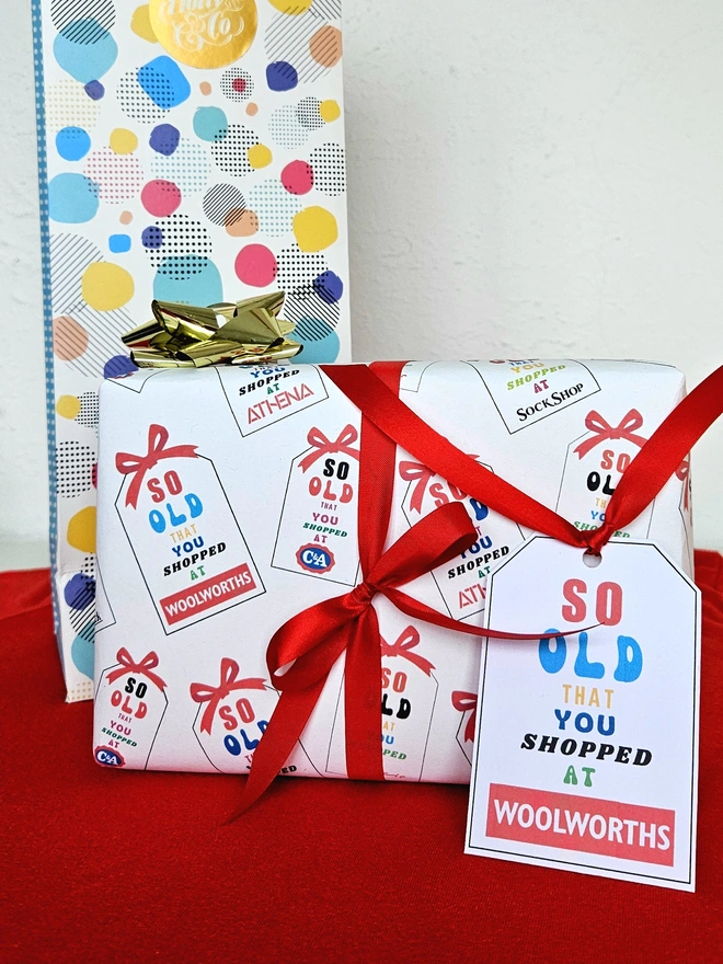A rectangular present gift wrapped in a design that says so old that you Christmas shopped at Woolworths, C&A, Sock Shop, Tie Rack, Athena on an illustrated white tag with a red bow. The text is written in bright  multi colours .