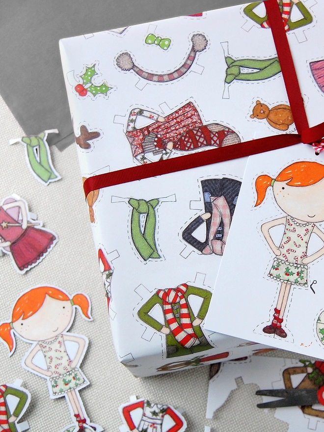 A gift wrapped in Christmas wrapping paper with illustrated outfits for the included paper doll tag.