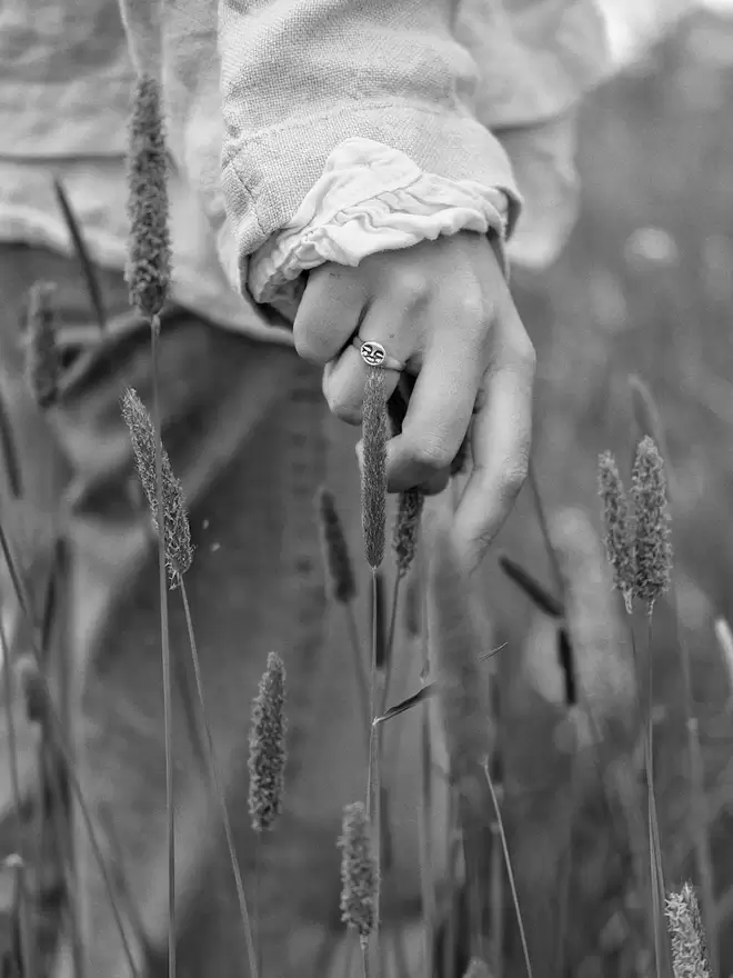 Black and white image of hand wearing a moon face ring, hand above long grasses and holding the top of grass