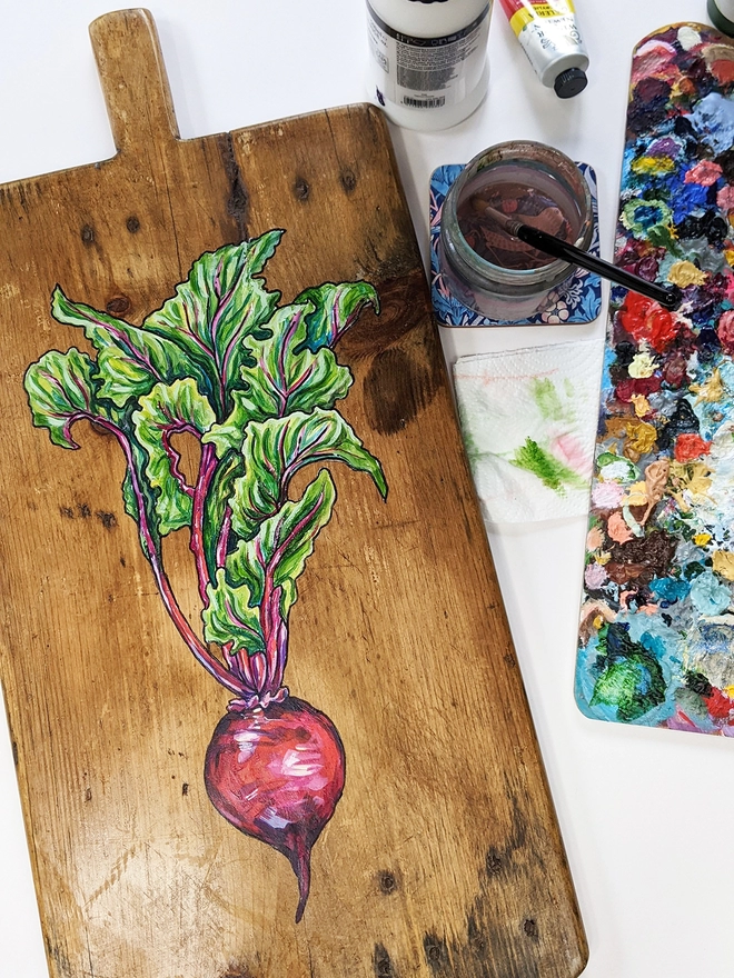 Wooden chopping board with handpainted design of a beetroot lying on a table next to artist's equipment