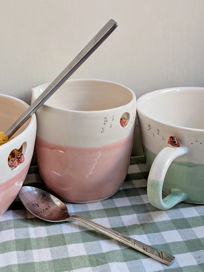 handmade ceramic white and pink jug with a tiny robin bird in a small cut out hole with bird prints and small musical notes hand painted next to a green robin cup and robin couple bowl