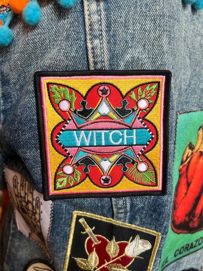 The patch has been embroidered onto a denim jacket, in between other patches. At the centre of the patch is the word “Witch” embroidered in white on a light blue strip. Surrounding this is a colourful, bold design which sits on a yellow background, with a thin pink inner border and a thick black outer border.