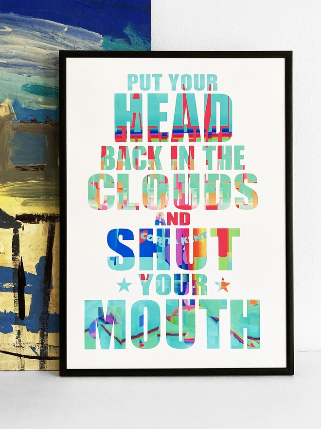 Framed multicoloured typographic print of a Julian Cope song, “put your head back in the clouds and shut your mouth”.  The print rests against a blue and yellow abstract painting.