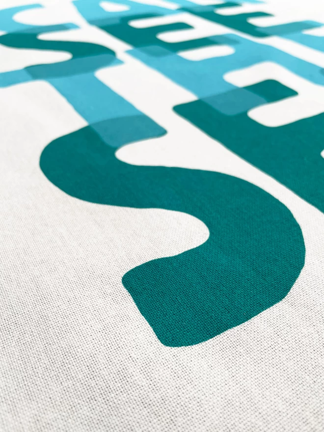 Close up of the turquoise ink printed lettering of I can see the sea, a big S is nearest with the cotton texture showing.