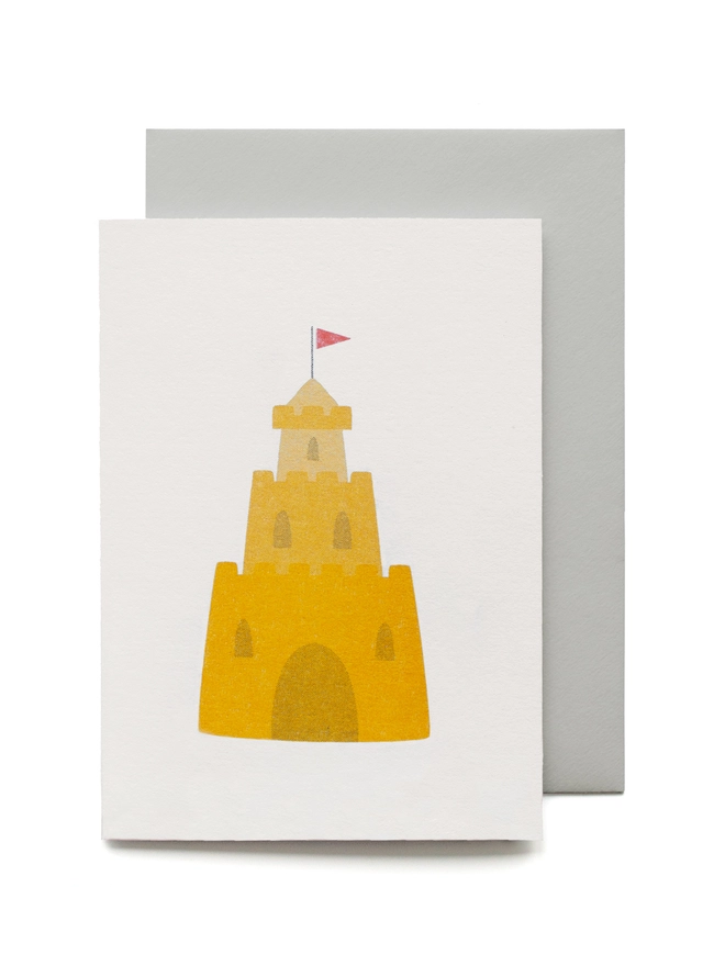 A yellow sandcastle is illustrated onto a small white card, a little red flag sits on the top tier of the castle. 
