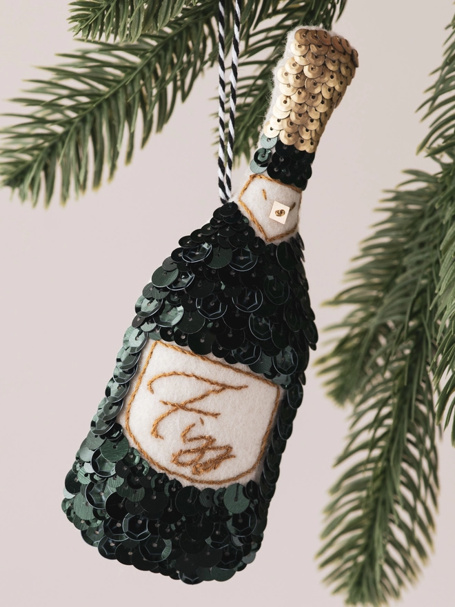 A green sequinned Champagne bottle ornament hanging on a tree