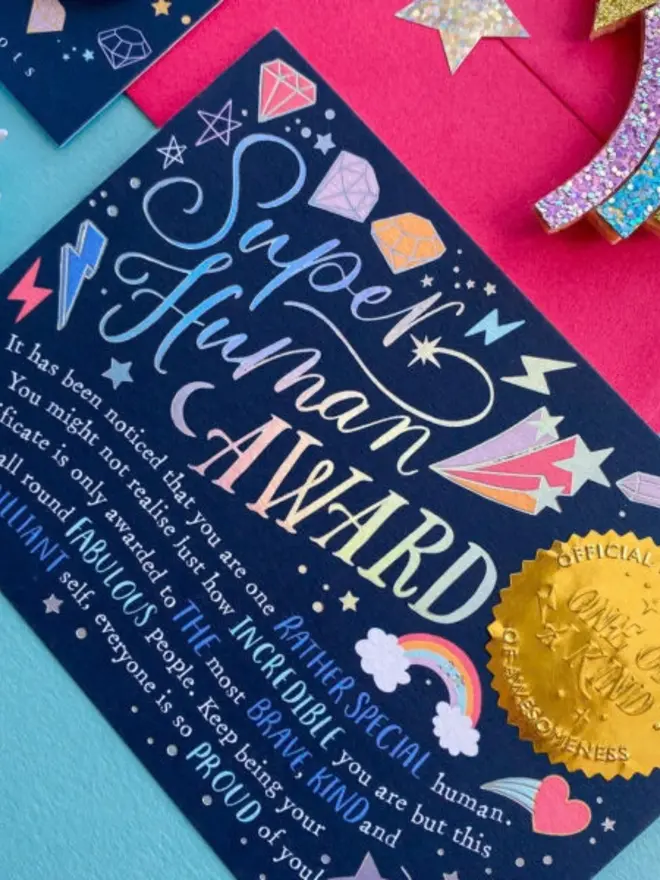 A version of the navy certificate with rainbow holographic foil lettering and an embossed gold seal. It rests on a pink envelope beside a glitter shooting star.