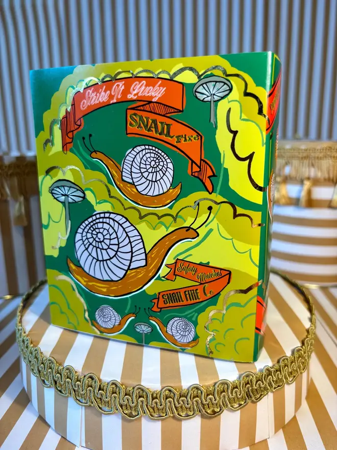 Snail Matchbox, a snail matchbox with a green design sitting on a decorated surface accompanied by other accessories.