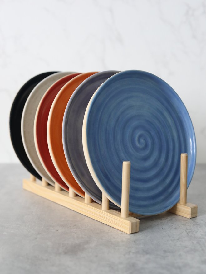 Six handmade ceramic side plates displayed upright in a natural wood plate rack, fanned out to reveal a colour palette of blue, mandarin orange, cherry red, speckled stone, and black, each showing a concentric swirl texture, against a white marble background