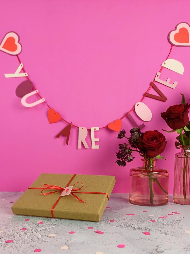 Paper garland with letters in various shades of red and pink spelling out the words you are loved. Garland ends are 3 layered pink hearts. 