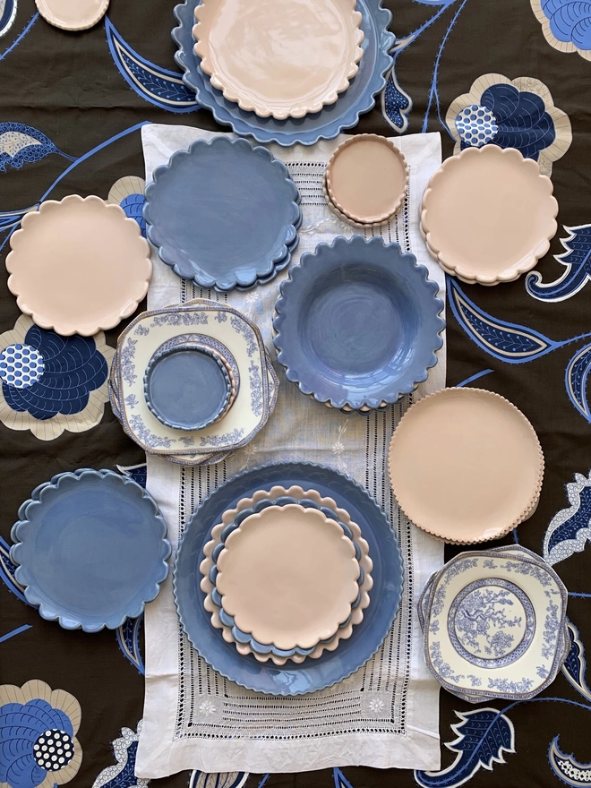floral fabric background with browns, blues and white with a variety of daisy edge and scaloped edge dinner and side plates scatered around. plates are periwinkle blue, white and blossom pink. some blue and white vintage cake plates shown also