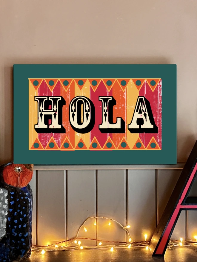Hola is hand painted onto reclaimed wooden sign with fairground lettering. with a teal frame and orange pink and yellow stripes with and orange and yellow zig zag at the top