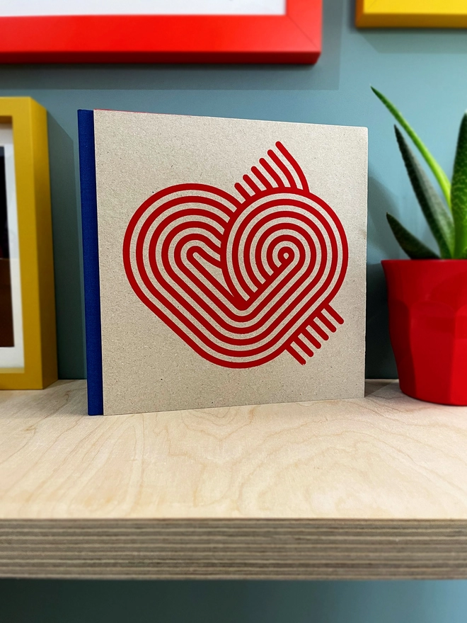 Stripy Heart design is screenprinted in magenta on a grey pasteboard sketchbook with a blue fabric spine, stood on a plywood shelf with hints of framed pictures around. the wall is duck egg blue, a plant glimpsed to the side.