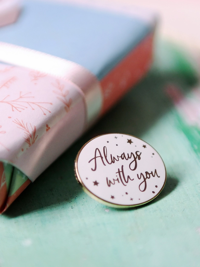 A white and gold enamel pin badge rests beside a wrapped gift. It has a subtle star design and the words "Always with you".