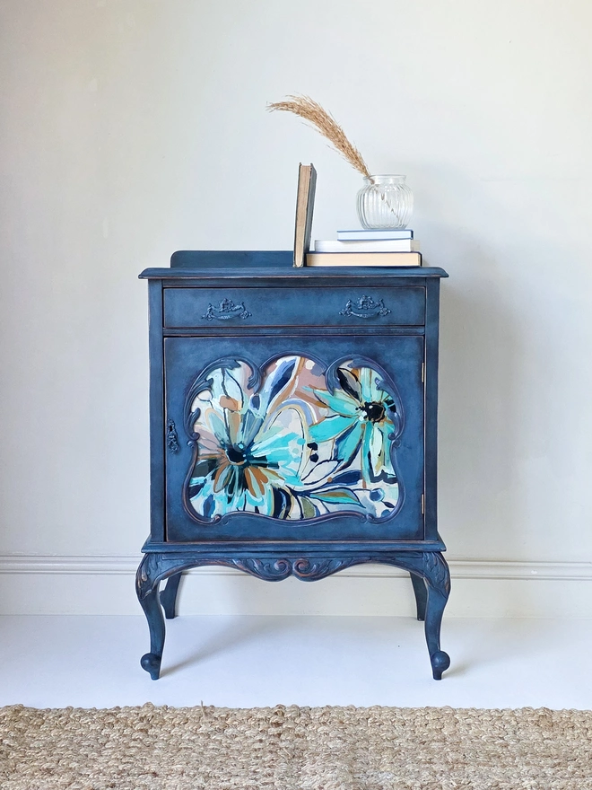 A french style bedside cabinet or drinks cabinet with beautiful carving especially on the legs. The frame has been painted a rich navy blue to contrast against a bespoke handpainted glass panel in the centre featuring daisies in turquoise, pink, white, and rusty orange. It is centre stage sitting in front of a rustic rattan rug, and sitting on top, vintage books, a reeded glass vase and pampas grass.