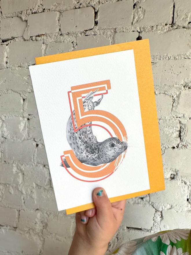 A white card featuring a big orange number 5 and a harbour seal illustration with a bright yellow envelope