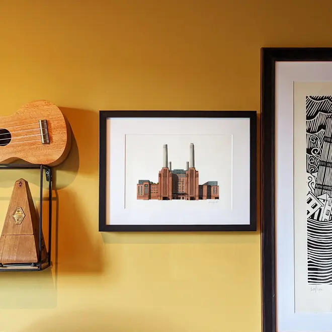 Battersea Power Station Giclée Print (Unframed), a print of Battersea Power Station sitting in a black frame against a mustard backdrop. It is joined by other framed prints. 