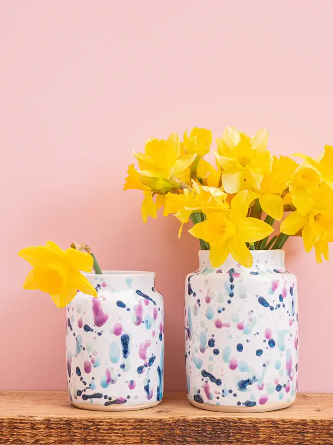 Sweetpea Medium Vase, a colourful vase with a collection of yellow flowers in it. It sits in front of a pink backdrop on a wooden surface. 