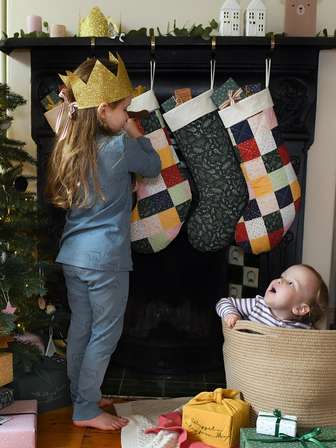 A young girl stands at a fireplace beside a Christmas tree with several patchwork stockings hanging up. She wears a golden crown and a toddler sitting in a woven basket looks at her.