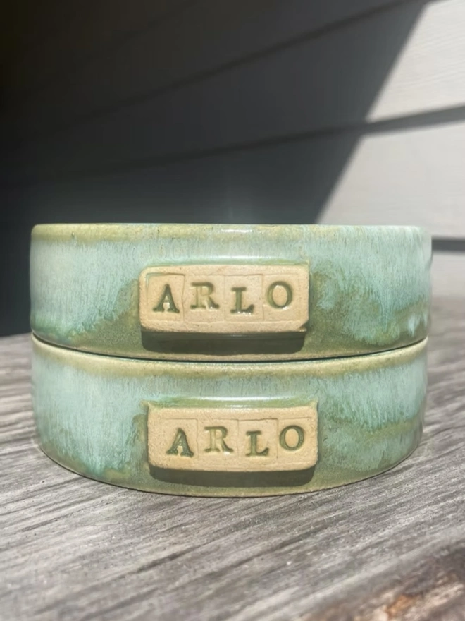 Personalised Handmade Ceramic Cat And Dog Bowls, green pet bowls stacked on top of each other that say 'Arlo' on a wooden surface. 