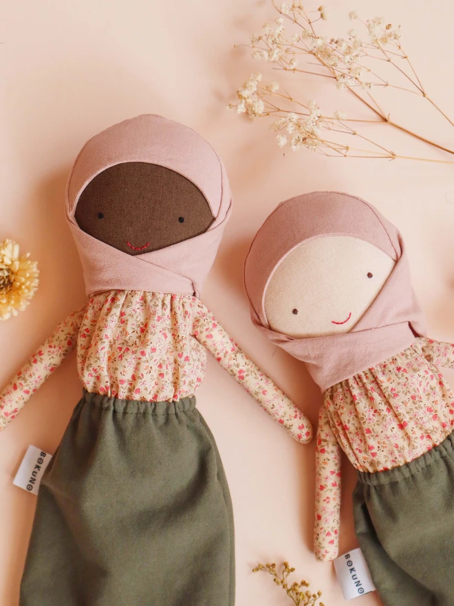 Two handmade cloth dolls wearing a blush pink hijab, a beige floral long-sleeve blouse, and an olive green skirt. The dolls have light and dark fabric skin, simple stitched eyes and a small red smile, with a BOKUNO label on the side. 