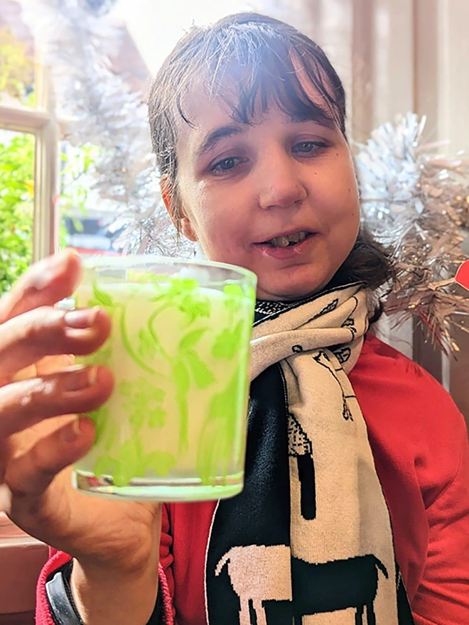 Artist holding laura's floral, wild fig & grape charity candle in a reusable glass with green illustrations