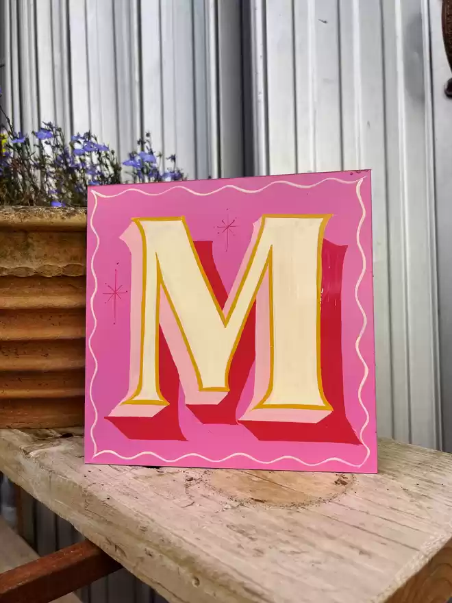 Bright and bold hand-painted panel with the letter 'M' in beige, golden yellow and various pink tones, propped up against a plant pot.