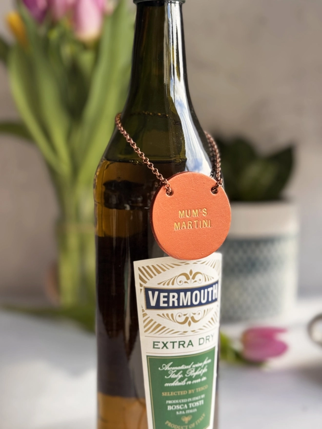 A dark glass bottle with a copper leather tag hanging around the neck by a delicate chain. The tag is hand-stamped with 'MUM'S MARTINI' in gold lettering, with pink tulips and a rustic background. A perfect personalised Mother’s Day gift or home bar accessory