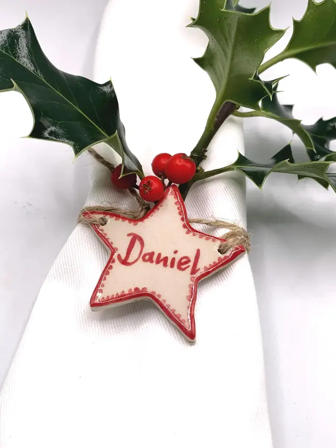 Personalised Ceramic Christmas Star Name Place Markers, a colourful ceramic start sitting on a white surface attached to a holly bush. 