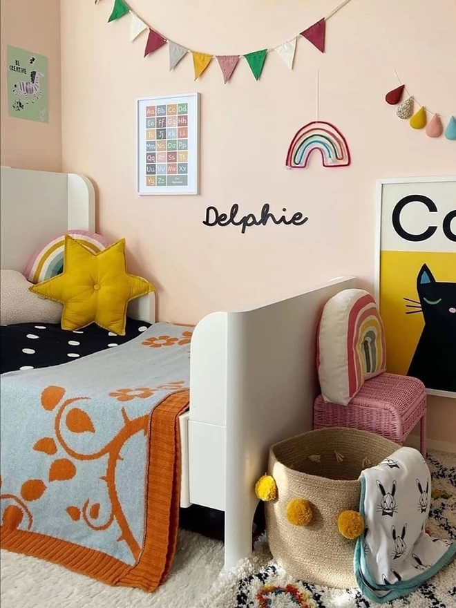 A bright and colourful kids room showing the rust briar rose blanket folded in half and draped over an extendable toddler bed. Bright pictures and bunting adorn the walls and a pom pom toy basket sits at the end of the bed.