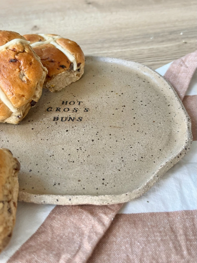 Speckled ceramic plate with the wording 'hot cross buns' in black text. The plate sits on a tablecloth serving up some delicious hot cross buns