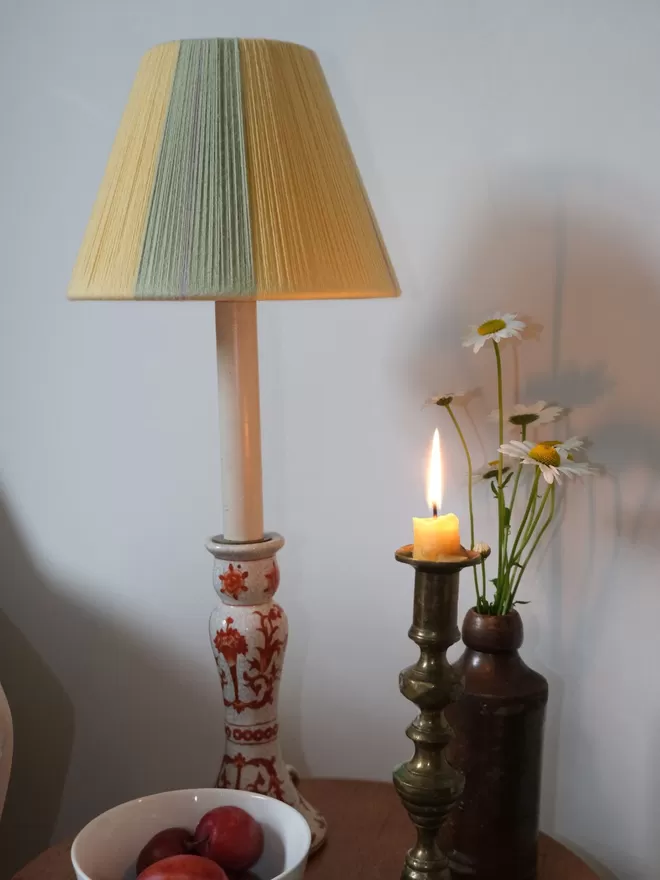Lifestyle image of Butter Yellow lampshades on a table lamp, placed on a table with a candela and flowers.