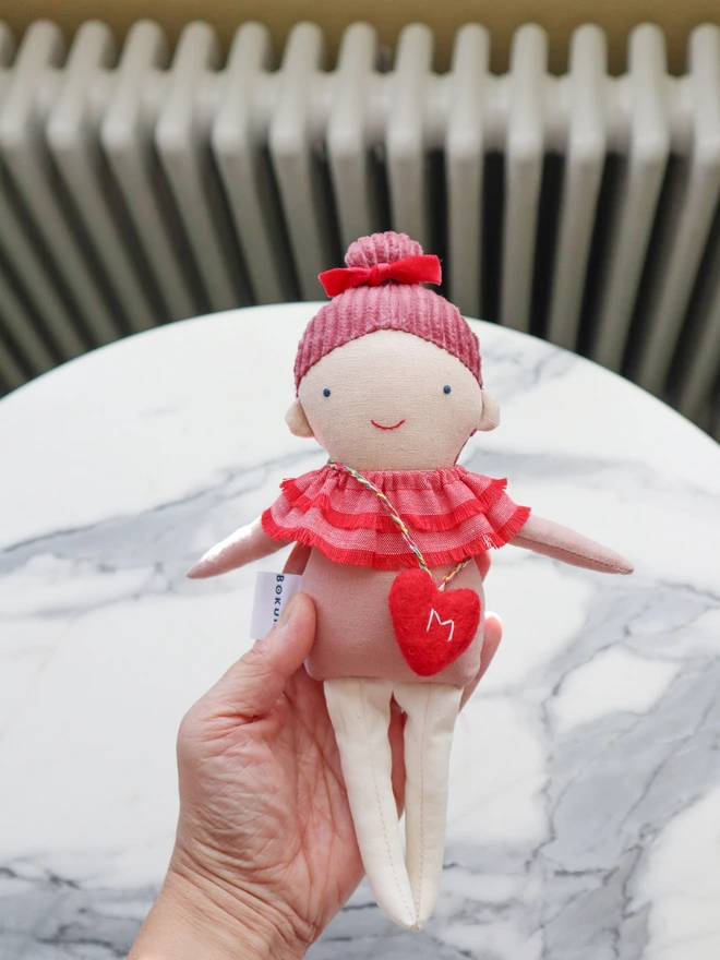Handmade cloth doll with a pink corduroy hair with a red velvet bow and a layered red ruffle collar and pink outfit, wearing a small red felt heart bag with the letter “M”