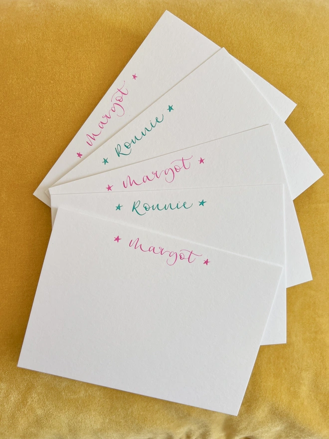 Five notecards in a fan with the names Margot and Robbie written on alternate cards in pink or green calligraphy, bookended by singluar stars