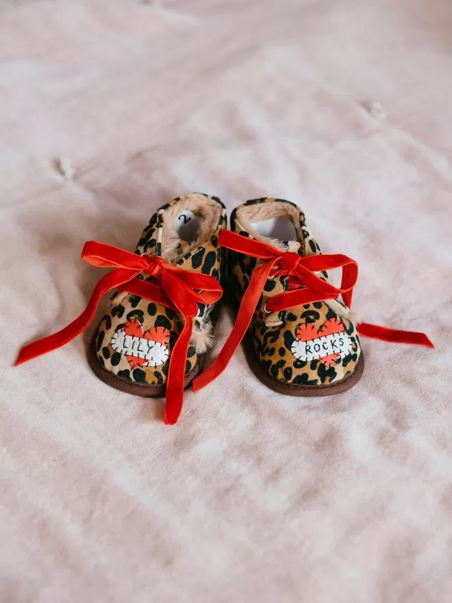 A pair of leopard print baby booties, lined with fur and with red velvet ribbon laces. A red heart with a white scroll adorns each booty. One says LILY, the other, ROCKS in black stitches