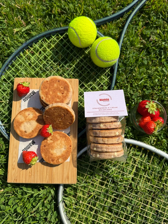 6 pack of strawberries and cream welsh cakes on a tennis racket, balls, and strawberries on the grass, with loose welsh cakes on a wooden board.