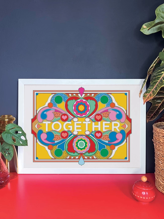 A landscape print on a white background with dark blue linework and green, blue red pink and gold sections. Across the centre of the image is written ‘TOGETHER’. The print is in a white frame on a red cabinet, and is resting against a dark grey wall. Next to the frame are two pot plants and a small red wooden pot.