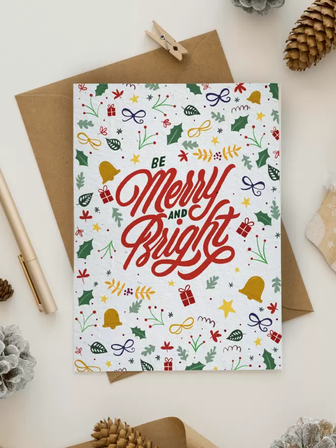 Christmas Card with 'Merry & Bright' in the centre with illustrations of presents, berries, holly leaves, bells and bows surrounded by pine cones, a pen and a small peg
