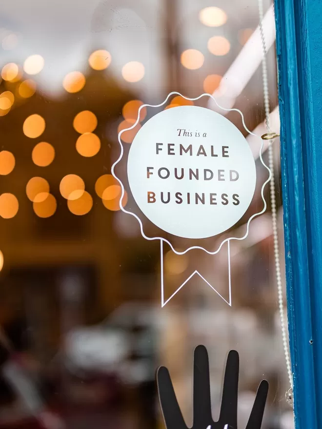Female Founded Business Rosette Sticker seen on a shop door with a blue frame and a black hand sticker below.