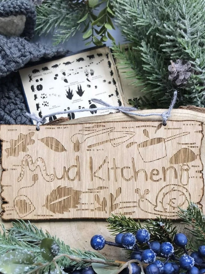 Personalised Wooden Mud Kitchen Sign, a personalised wooden mud kitchen sign sitting on some foliage outside. 