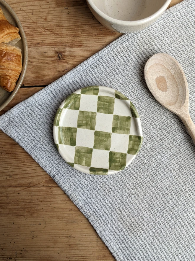 A green and white checkerboard design spoon rest, on a wooden table, next to a wooden spoon