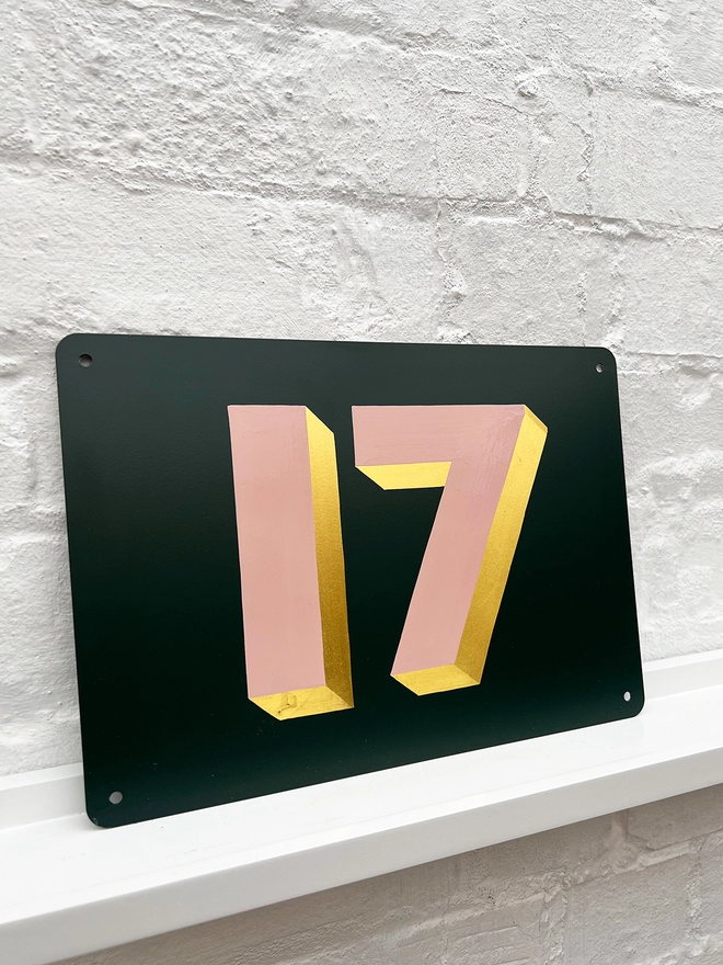 Hand painted dusky pink and gold leaf house number 17 on an anthracite grey metal plaque against a white brick wall. 
